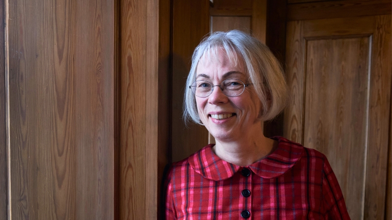 En vit leende kvinna, 66 år. Grå page, bär ovala glasögon och en klänning med rundad krage med röda och blå ränder i olika nyanser. Står vid ett fönster.