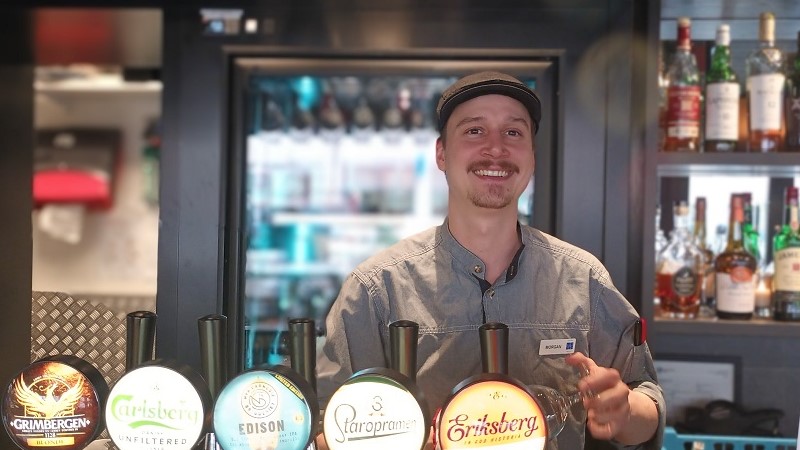 En ung man i farfarskeps torkar ett ölglas i en bar med flera ölkranar. Han har brun mustasch och lätt hakskägg.