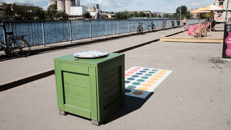 En bred gångväg löper längs vattnet en solig dag. I förgrunden står en grön kubformad låda bredvid ett färgglatt twisterspel målat på marken, och längre bort syns cyklar, solstolar och en uteservering vid kajkanten.