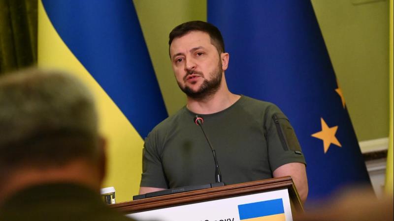 Medelålders vit man med snaggat brunt hår, helskägg och mustasch. Har militärgrön T-shirt. Står vid talarpulpet. Bakgrund- en Ukrainsk flagga i blått och gult. 