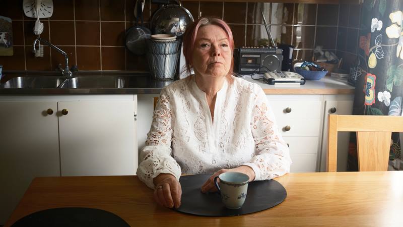 En vit kvinna 70 år. Sitter vid ett köksbord i ljust trä med kaffekopp framför sig. Rosa hår, bär vit mönstrad spetsblus.