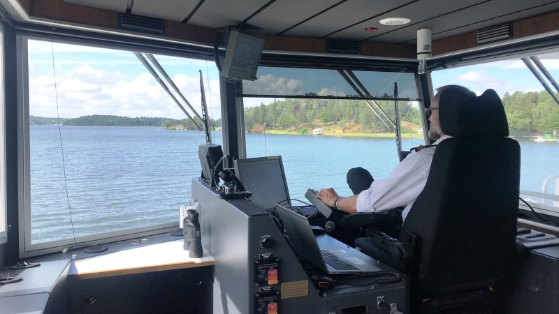 I styrhytten med stora fönster ser man kapten i vit skjorta snett bakifrån i stor fotölj. Utsikt över vatten, till höger land i sommargrönska., 