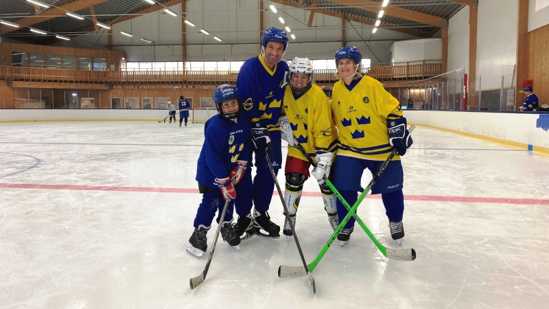 Tre vuxna och ett barn i ishockeymundering med klubbor på isrink.