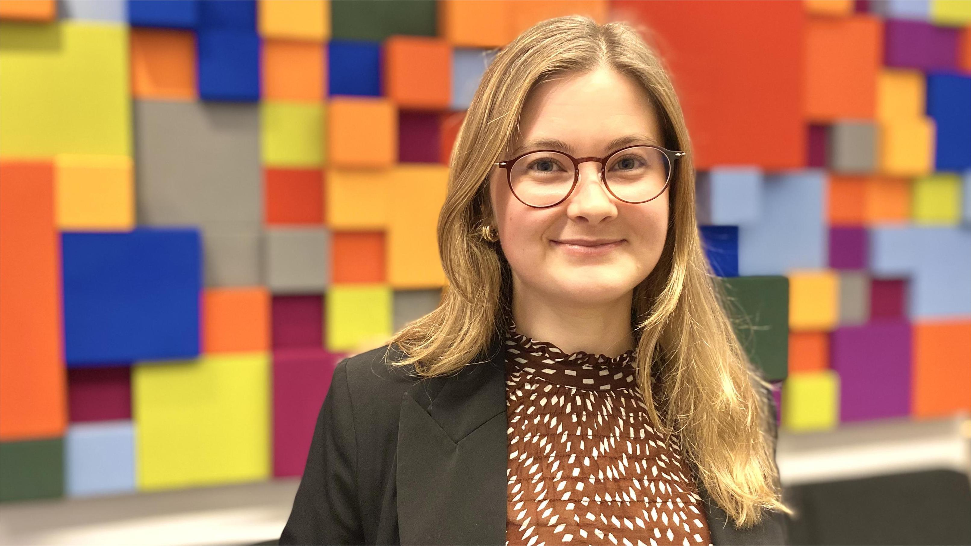Johanna Carlsson verksamhetsspecialist pensionsmyndigheten. Ung vit kvinna med rågblont långt hår, och runda glasögon. Svart kavaj med brun vitprickig blus under. Står framför ett väggkonstverk av färgglada kuber