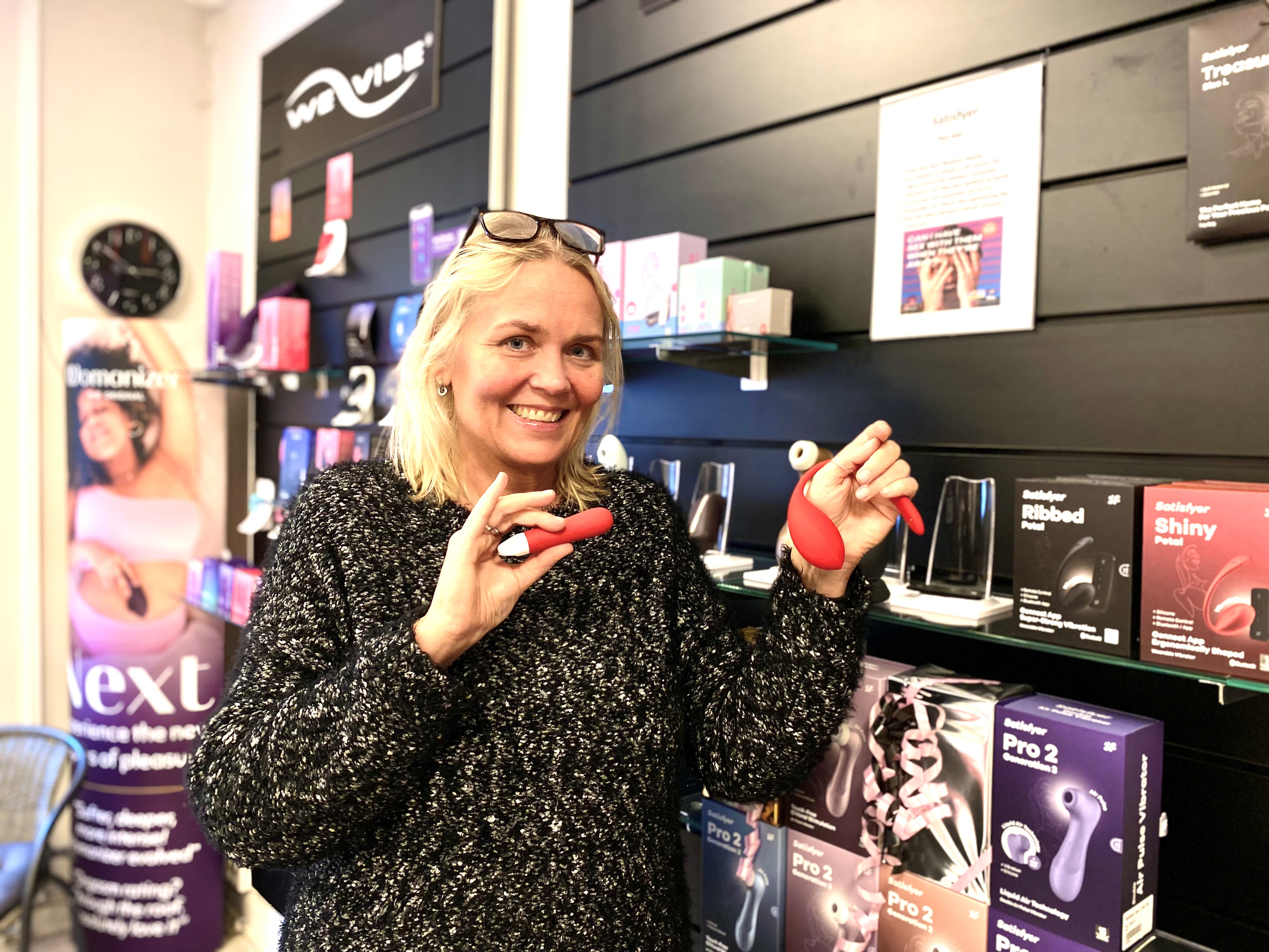 En vit blond medelålders kvinna i svartglittrig tröja, håller i två mörkrosa små sexleksaker- vibratorer. Står i butik med sexprodukter.