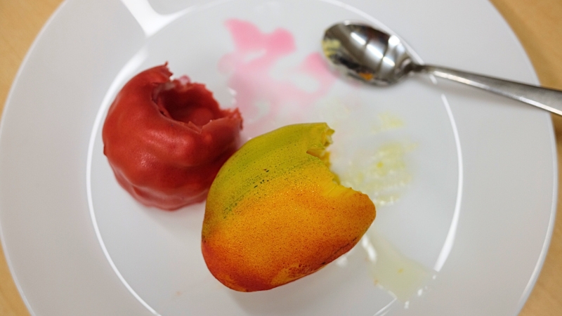 En röd bakelse i formen av ett hallon ligger på en vit porslinstallrik bredvid en bakelse i formen av en mango. Båda bakelserna är halvätna, och på tallriken ligger även en sked.