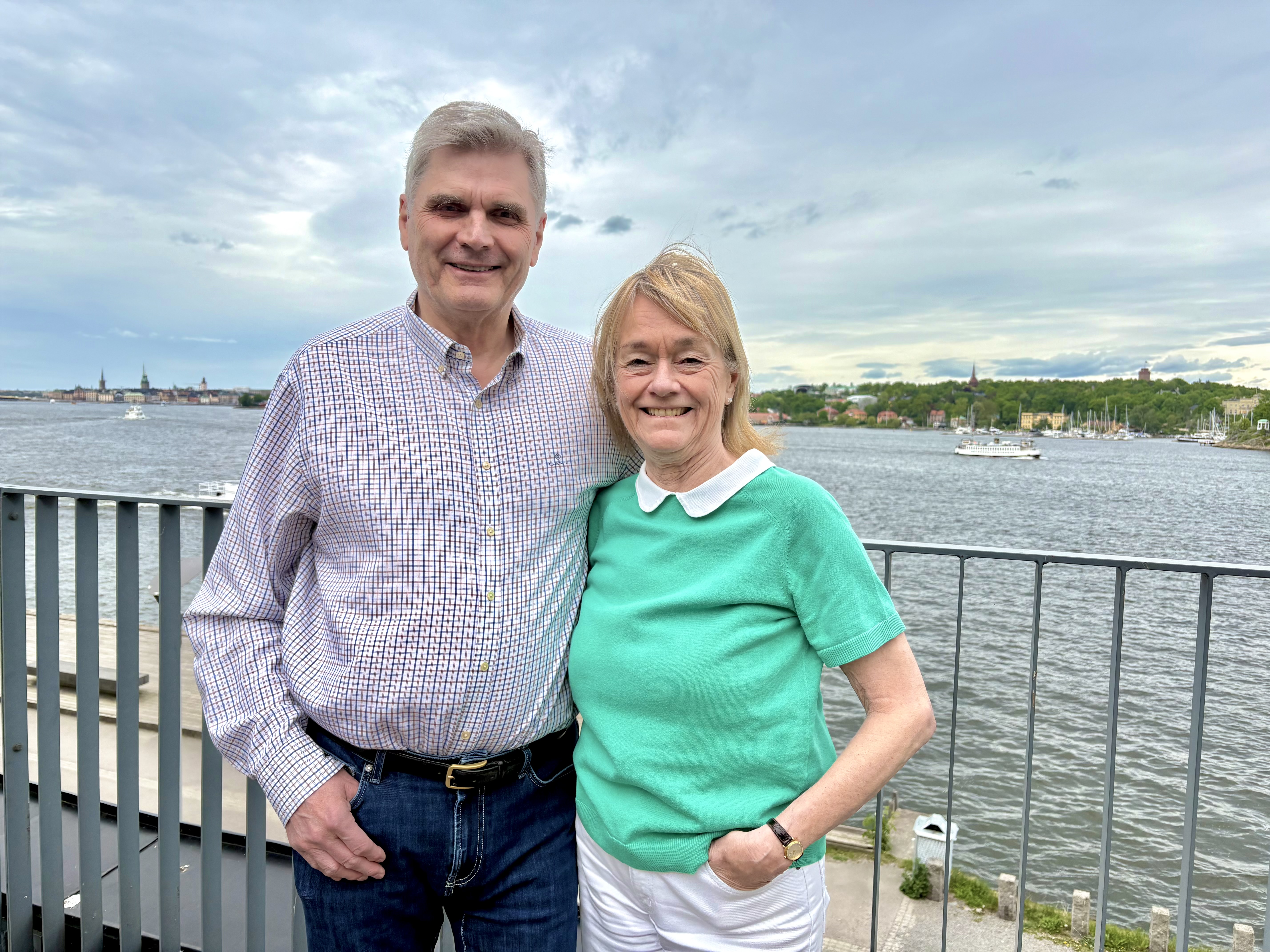 70-årigt vitt par håller om varandra vid ett terassräcke med utsikt över Stockholms inlopp. Han har kort grått hår, småvitrutig skjorta och blåjeans, hon har ljusgrön t-shirt och vita jeans.