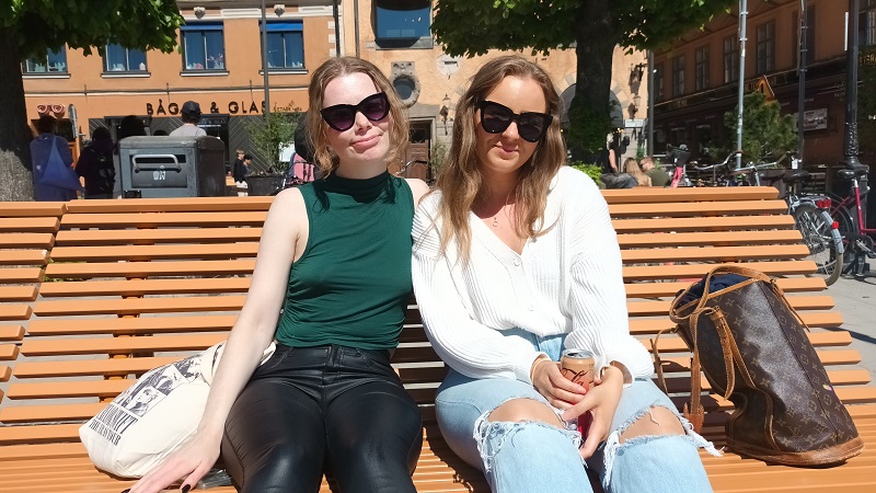 Två unga kvinnor, båda med mörkblont långt hår och solglasögon sitter i solen på en stor orange träbänk med bakåtlutande ryggstöd.