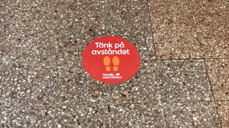 Rött stort klistermärke på stengolv i affär, med text, tänk på avståndet, handla med hänsyn