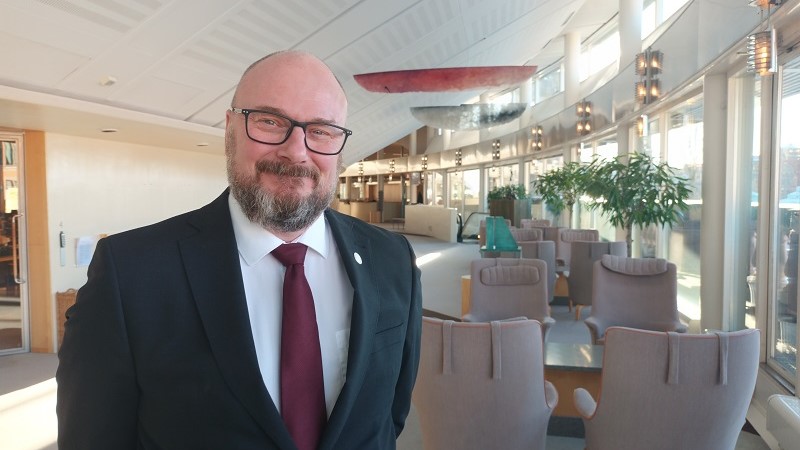 En vit man med kal skalle, glasögon, grått skägg och svart kavaj med röd slips står i en ljus hall i riksdagen.
