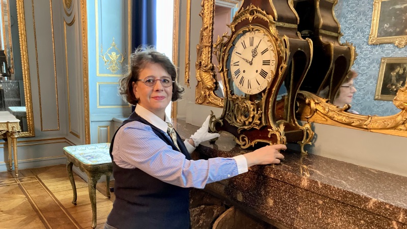 Medelålders vit mörkhårig kvinna står vid gammaldags bordsur i guld. Är på slott