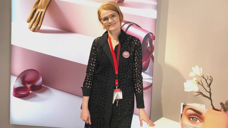 Svartklädd leende kvinna med kort, ljusrött hår i snedbena och ljusa pilotglasögon i elegant butiksmiljö.