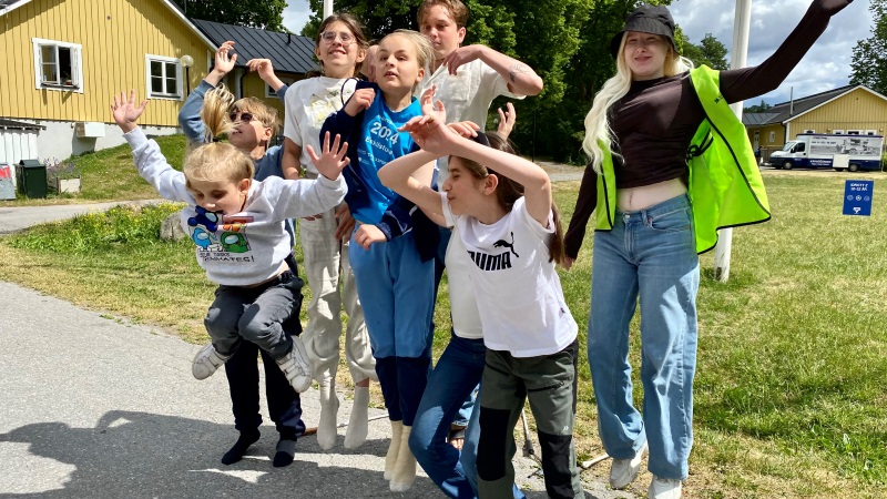 Några barn i 10-årsåldern hoppar upp med armarna uppsträckta. De står på gräsmatta. Sommar.