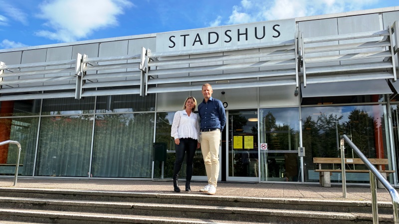 En kvinna och en man står bredvid varandra på en stor trappavsats utanför Lidingö stadshusentré. De är båda vita, medelålders. Hon har vit spetsblus och svarta skinnbyxor, han har mörkblå skjorta och beiga byxor. 