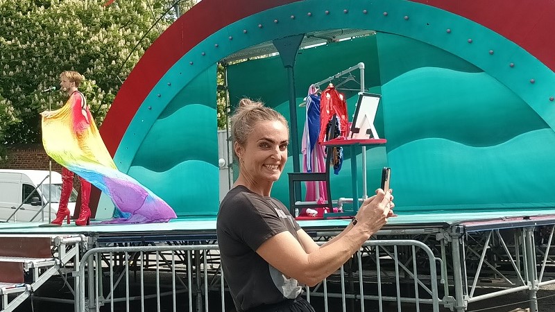 En kvinna med blont hår uppsatt i knut står framför en scen med grön dekor och fotar något utanför bild med mobilen. På scenen syns en man i regnbågsfärgad cape och höga röda lackstövlar.