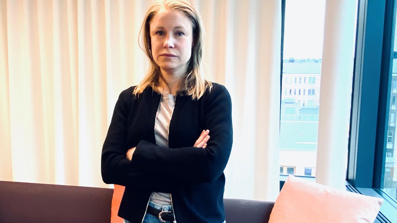 Vit medelålderskvinna, axellångt blont hår i mittbena. Svart kofta, blåjeans. Armarna i kors. Står vid fönster med beige gardin, utsikt stadshus.
