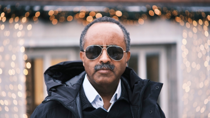 En svart man, 65, år, med svarta pilotglasögon. Han har mustasch och snaggat hår , gråsprängt. Han bär en vit småprickig skjorta, och över det en mörk tröja och en öppen svart jacka.