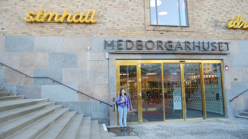 utomhusentrén på Forsgrenska badet, glasdörrar på sandfärgat stenhus, kvinna står framför.