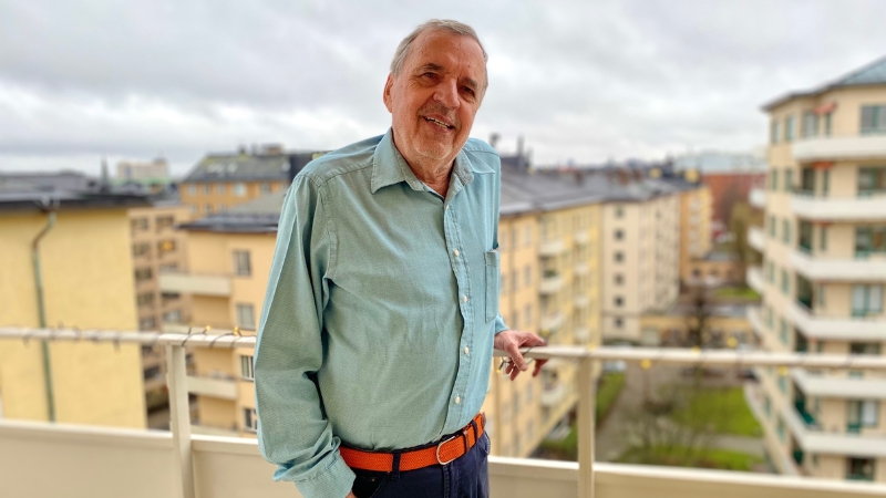Vit 80 årig man med kort grått hår, ljusblå jeansskjorta, mörkblå byxor och orange skärp. Står på balkong med vy över statsgata kantad av höga gula funkishus. Parkområde syns också.