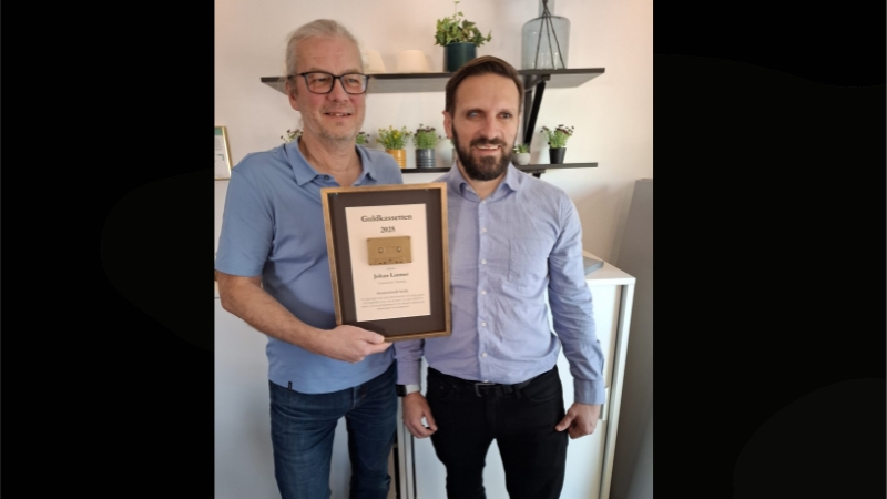 Två vita män stor bredvid varann i en konferenslokal. Han till vänster har ljusblå t-tröja, grått hår i tofs, glasögon och skäggstubb. Håller i ett guldinramat diplom. Mannen till höger har ljusblå skjorta, brunt kort hår och mörkt kort skägg.