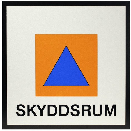 Skylt för Skyddsrum. På en vit botten en orange kvadrat, inuti den en mörkblå uppåtriktad triangel. Svart text i underkant: SKYDDSRUM