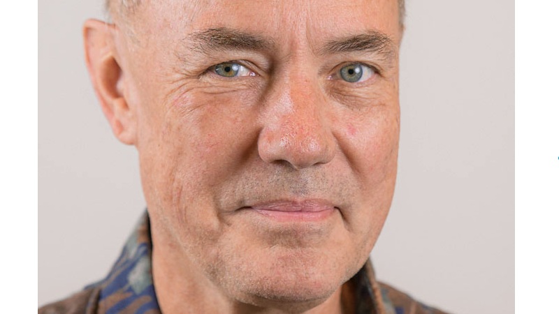 Närbild ansikte, vit 65 årig man, renrakad, blågrå ögon. Kort vitt snaggat hår, buskiga bruna ögonbryn. Brunmelerad jackkrage syns.