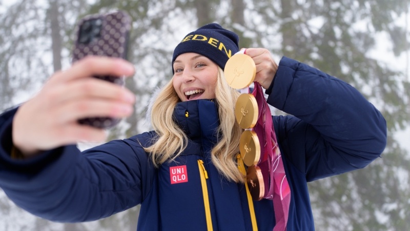 En blond vit kvinna i 25-årsåldern tar en selfie med sin telefon. Hon bär en mörkblå mössa och en mörkblå jacka och visar upp fyra medaljer för kameran, Tre guld och ett brons. 