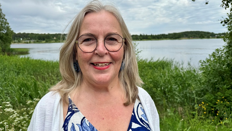 En vit 67-årig leende kvinna med axellångt grått hår, och runda mörkbågade glasögon. Vit kofta och blåblommig klänning. Står framför en sjö med grönskande vass och skog runt. Sommar.