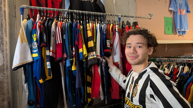 22-årige Noel Lindgren står inomhus framför en klädställning fylld med färgglada fotbollströjor. Noel har solglasögon i pannan som håller tillbaka hans halvlånga lockiga hår. Han bär en svartvitrandig fotbollströja.