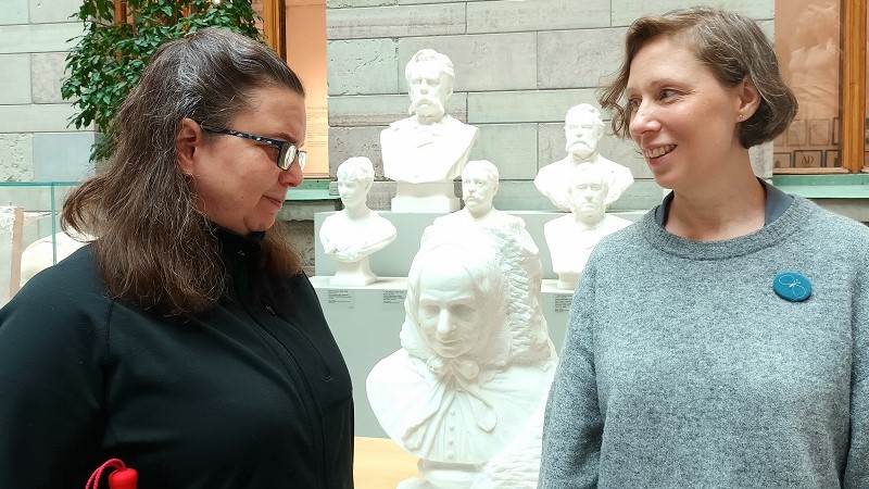 Två kvinnor står på varsin sida om en marmorskulptur, byst av Fredrika Bremer, med ansiktena mot varandra. Anne Mondotter till vänster, vit med glasögon och mörkt hår med grå stråk, i en uppsättning och fall över axlarna. Till höger Sara Borgegård Älgå, vit med en ljusbrun pagefrisyr. I bakgrunden finns fler marmorskulpturer.