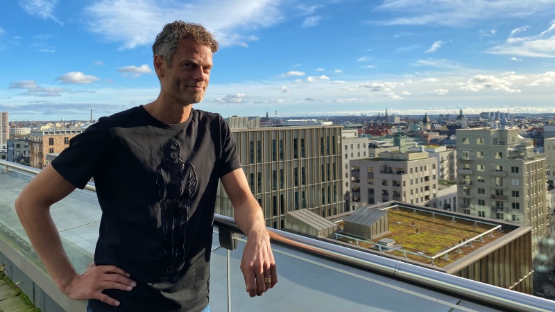 Vit medelålders man med kort grått hår, och svart t-shirt. Står lutad vid ett balkongräcke med utsikt över Stockholms takåsar.