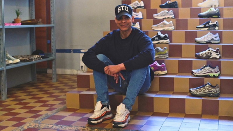 Isse, en man med brun hy i yngre medelåldern sitter på en kaklad trappa. Har svart keps med bokstäverna STU i vitt, mörk tröja och ljusblå jeans. På trappan bakom honom står olika skomodeller uppställda.