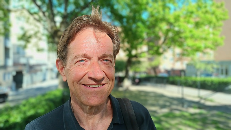 En medelålders vit man med bakåtkammat vågigt brunt hår och blå ögon står i en parkmiljö.