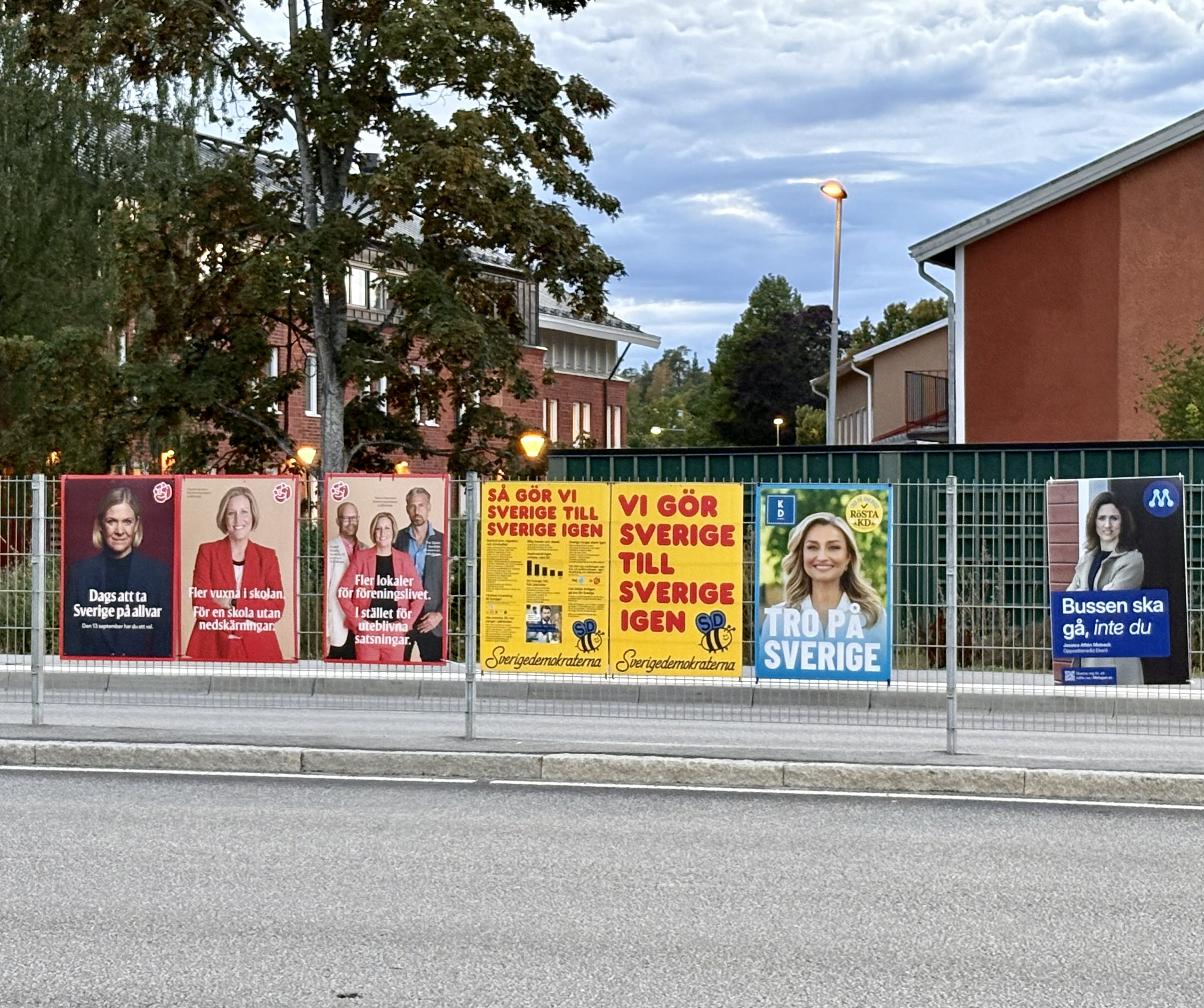Flera valskyltar från de politiska riksdagspartierna hänger på ett stålstaket i mitten av tvåfilig väg. Bostadshus i bakgrunden.