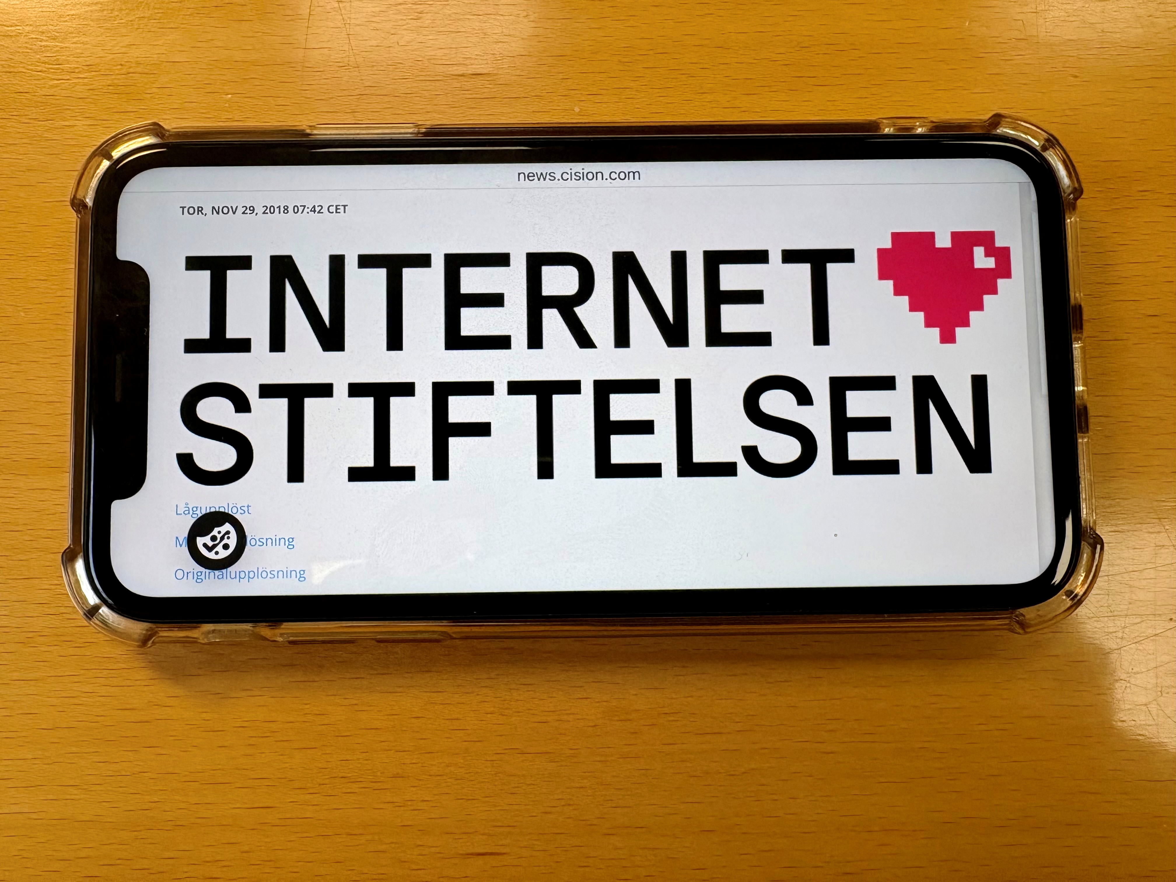 : Bild på mobil på ljust träbord. På skärmen svart text på vit botten: Internetstiftelsen. Ett rosa hjärta bredvid texten