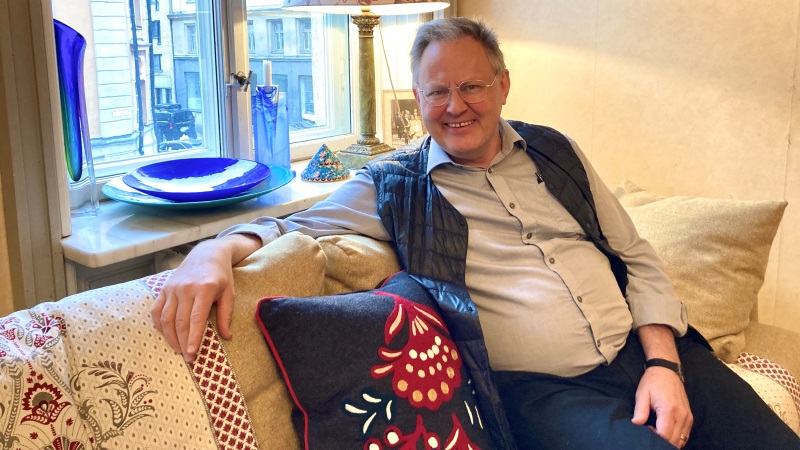 En vit lite rundlagd man med grått hår och glasögon sitter i en soffa framför ett fönster med glaskonst.