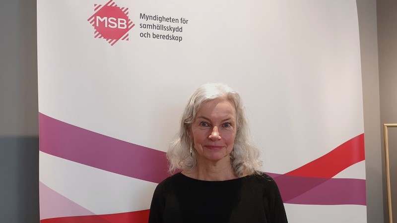En vit medelålders kvinna med axellångt vågigt vitt hår står framför en banderoll med logotypen för MSB, i form av två överlappande fyrkanter i rött och vitt.