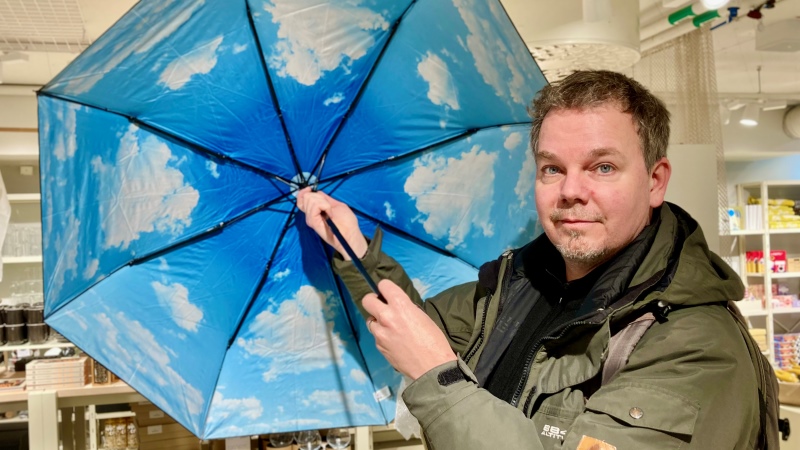 En vit medelåldersman med kort brunt hår, står med ett uppspänt paraply vars insida är tryckt med blå himmel med moln. Han står i en butik.