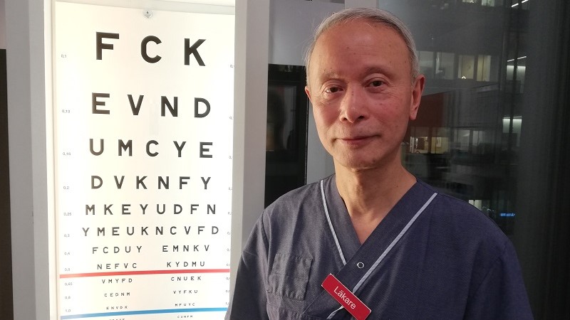 En asiatisk man med tunt grånat hår står vid en syntavla med gradvis mindre bokstäver. På hans blå arbetsklädsel sitter en skylt med texten LÄKARE.