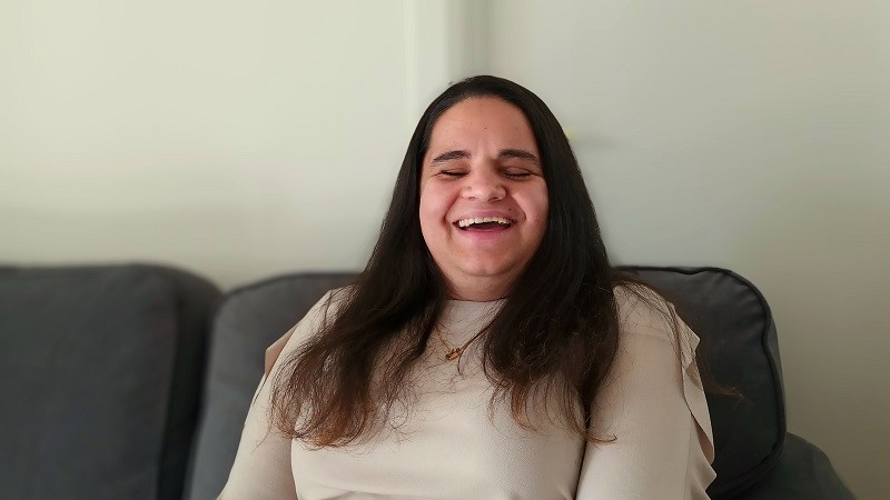 Sandra Ristic, en kvinna i 30-årsåldern med mörkt långt hår sitter i en grå soffa och ler stort.