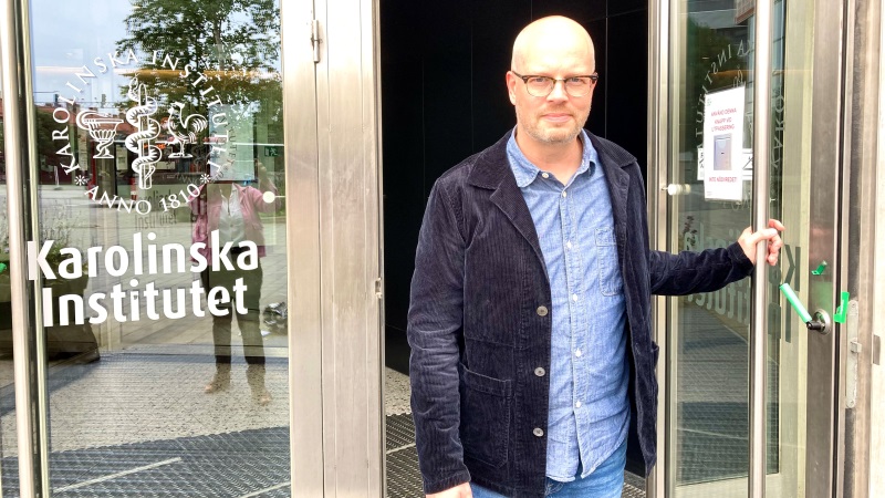 Vit medelålders skallig man i mörkblå manchesterkavaj håller upp en glasad entrédörr i högt hus.