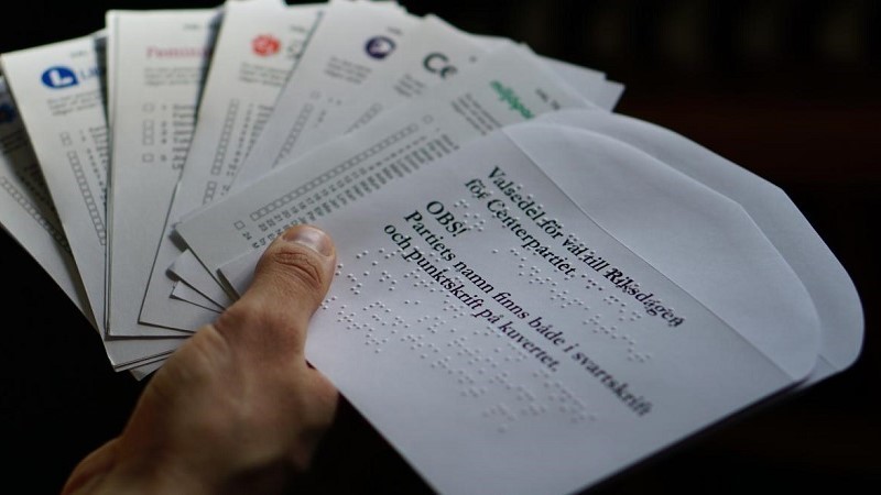 En hand som håller flera olika partiers valsedlar, och ett kuvert med punktskrift och svartskrift som lyder: "Valsedlar för val till riksdagen för Centerpartiet. OBS! Partiets namn finns både i svartskrift och punktskrift på kuvertet."