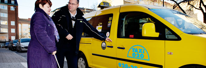 En kvinna i lila kappa med vit käpp visas till en gul färdtjänsttaxi av en chaufför i svart.