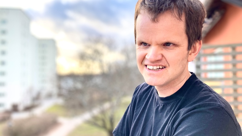 yngre man med kort brunt hår, svart t-shirt, står på balkong. Vita höghus och himmel i bakgrund.