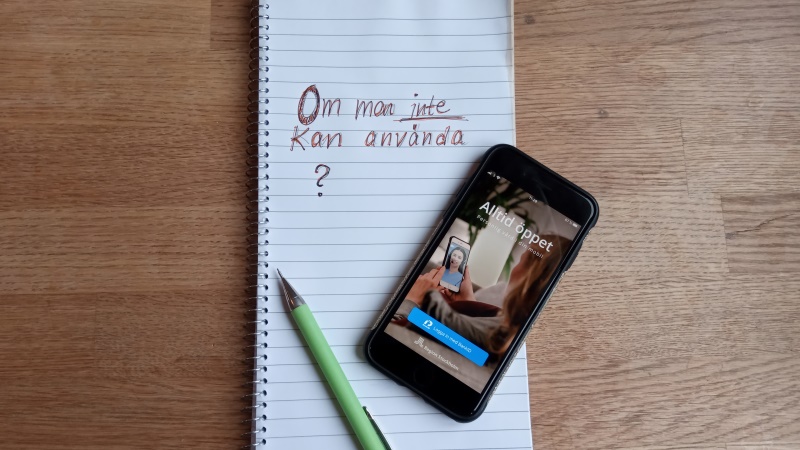 På träbord ligger vitt block, grön penna och mobiltelefon. Appen Alltid Öppet syns i displayen