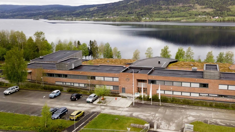 Ett lågt, avlångt tegelhus med mörkt tak ligger nära en stilla sjö. Framför byggnaden finns en parkeringsplats med några bilar. Bakom huset syns träd och långt bort på andra sidan sjön höga, gröna skogskläddaberg.
