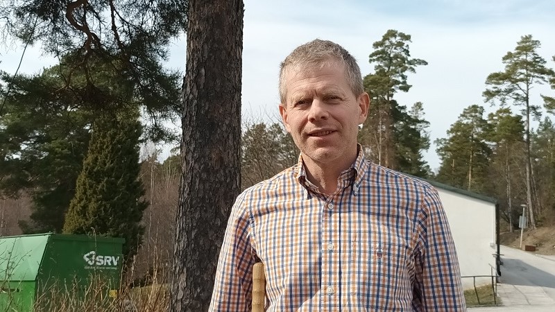 En medelålders vit man med kort gråsprängt hår och smårutig skjorta i blått, brunt och vitt, utomhus bredvid en tall.