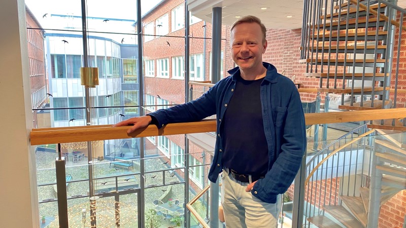 En medelålders  rödhårig vit  man står i glasat trapphus. Mörkblå skjorta och jeans.