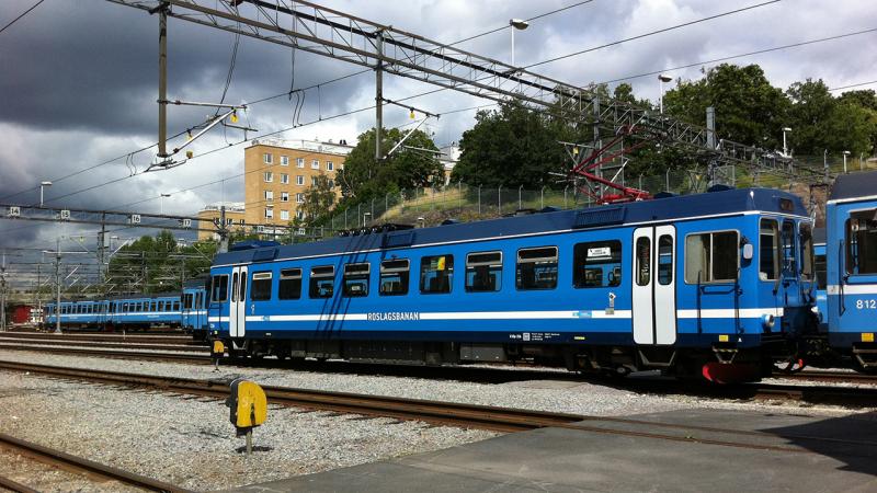 Ett blått tåg på räls, roslagsbanan. Kör i landskap med träd.