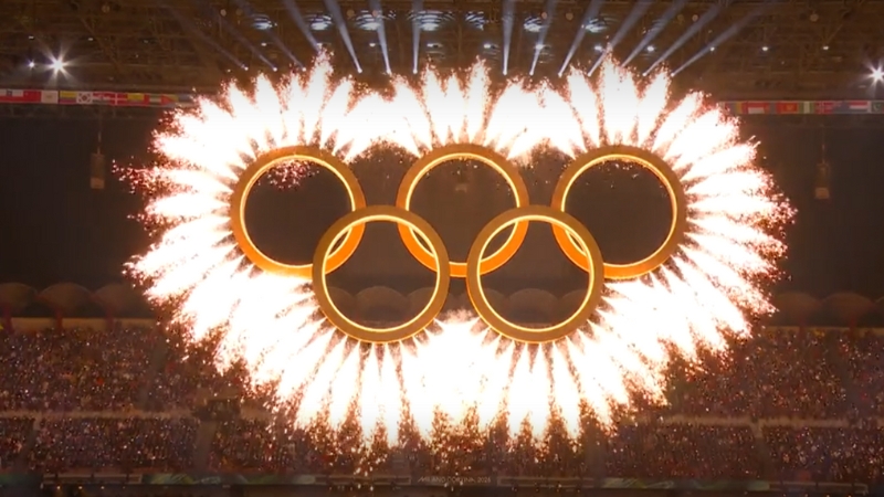 Bild från OS-invigningen där de fem olympiska ringarna i ett guldmaterial svävar högt över marken med publik i bakgrunden. Från ringarna sprutar gnistor som från fyrverkerier.